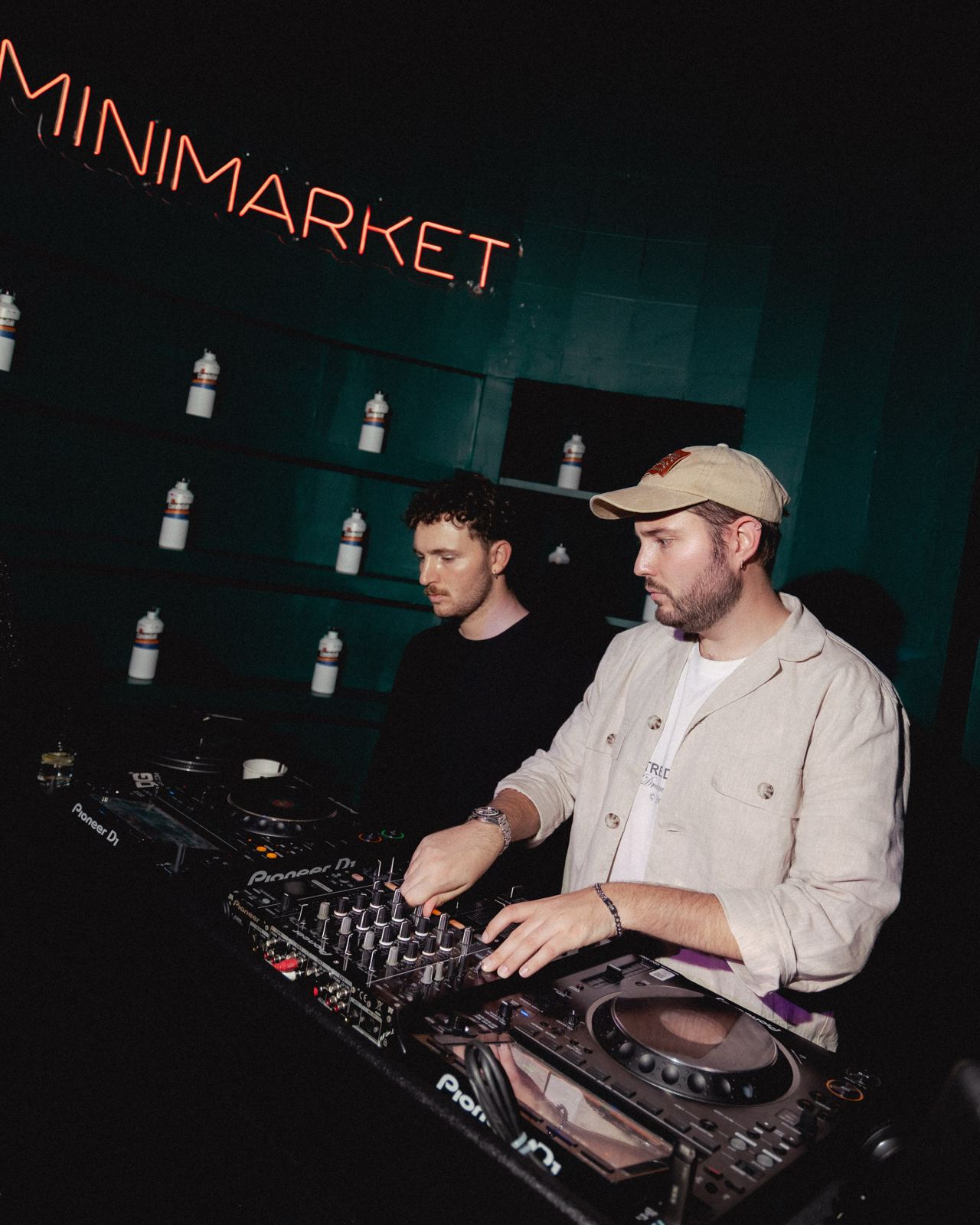 Due DJ in console sotto l'insegna al neon Minimarket