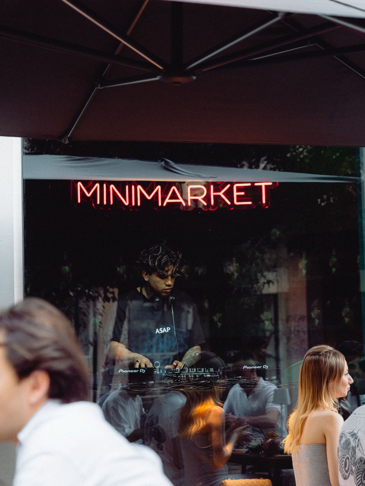 L'ingresso di Minimarket la sera, con l'insegna al neon accesa e gente che chiacchiera davanti